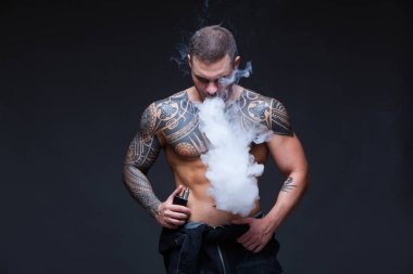 Vaper. Dövmeli bir kaslı çıplak gövde adamla koyu arka plan üzerinde elektronik sigara duman