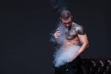 Vaper. Dövmeli bir kaslı çıplak gövde adamla koyu arka plan üzerinde elektronik sigara duman