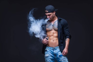 Vaper. Adam mavi kot pantolon giymiş, siyah gömlek ve siyah beyzbol şapkası dövmeler ile koyu arka plan üzerinde elektronik sigara duman.
