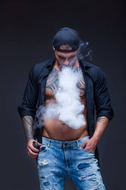 Vaper. Adam mavi kot pantolon giymiş, siyah gömlek ve siyah beyzbol şapkası dövmeler ile koyu arka plan üzerinde elektronik sigara duman