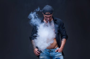 Vaper. Adam mavi kot pantolon giymiş, siyah gömlek ve siyah beyzbol şapkası dövmeler ile koyu arka plan üzerinde elektronik sigara duman