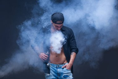 Vaper. Adam mavi kot pantolon giymiş, siyah gömlek ve siyah beyzbol şapkası dövmeler ile koyu arka plan üzerinde elektronik sigara duman