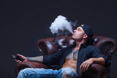 Vaper. Dövmeli adam koyu arka plan üzerinde elektronik sigara duman deri kanepede oturuyor.