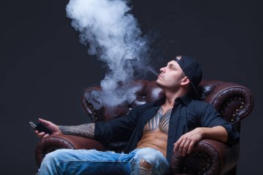 Vaper. Dövmeli adam koyu arka plan üzerinde elektronik sigara duman deri kanepede oturuyor.