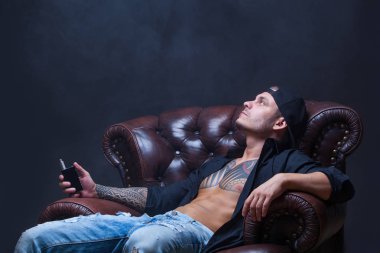 Vaper. Dövmeli adam koyu arka plan üzerinde elektronik sigara duman deri kanepede oturuyor.