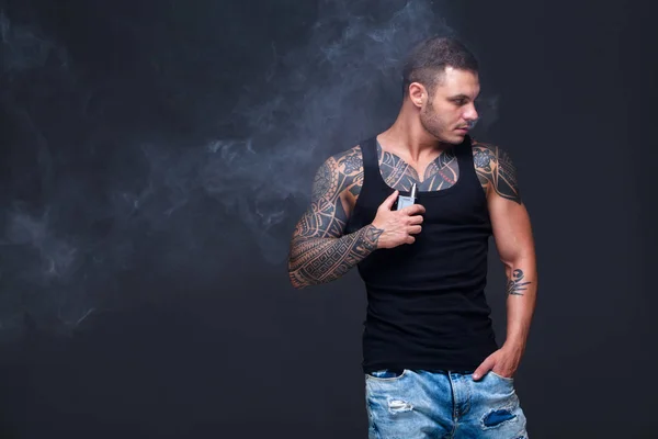 Vaper. El hombre con un torso muscular con tatuajes fuma un cigarrillo electrónico en el fondo oscuro — Foto de Stock
