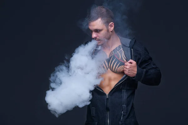 Vaper. Mannen med en muskulös nakna torso med tatueringar röka en elektronisk cigarett på den mörka bakgrunden — Stockfoto