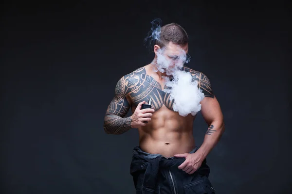 Vaper. De man met een gespierde naakte torso met tatoeages roken een elektronische sigaret op de donkere achtergrond — Stockfoto