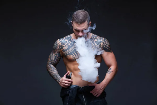 Vaper. De man met een gespierde naakte torso met tatoeages roken een elektronische sigaret op de donkere achtergrond — Stockfoto