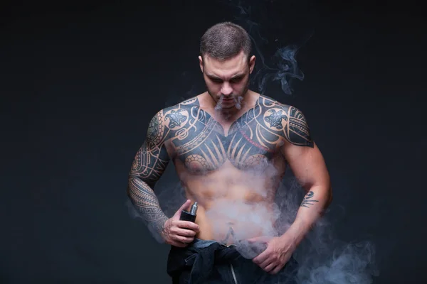 Vaper. El hombre con un torso desnudo muscular con tatuajes fuma un cigarrillo electrónico en el fondo oscuro —  Fotos de Stock