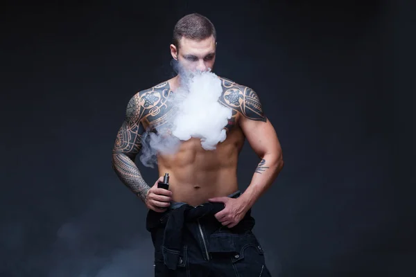 Vaper. El hombre con un torso desnudo muscular con tatuajes fuma un cigarrillo electrónico en el fondo oscuro — Foto de Stock