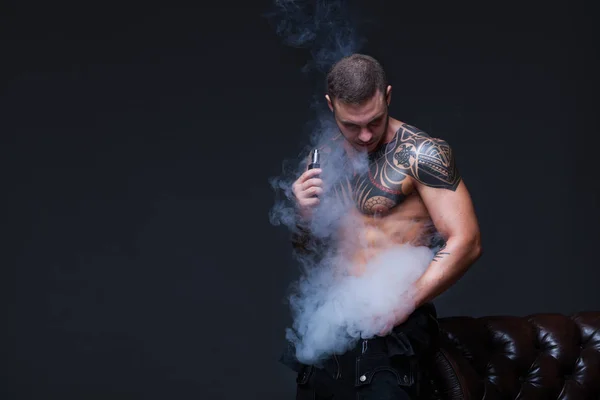 Vaper. El hombre con un torso desnudo muscular con tatuajes fuma un cigarrillo electrónico en el fondo oscuro —  Fotos de Stock