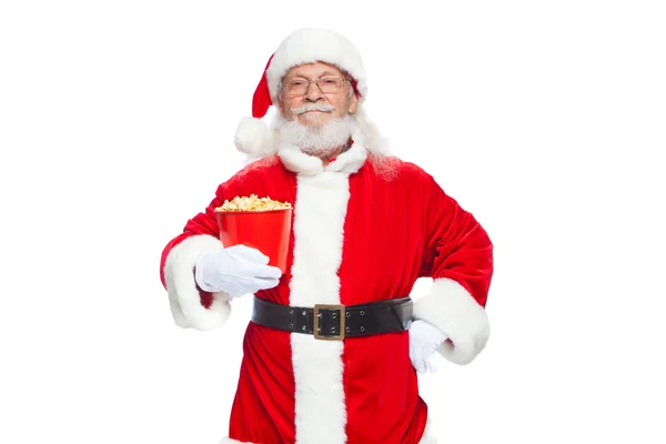 Natale. Sorridente, gentile Babbo Natale in guanti bianchi tiene un secchio rosso con popcorn con una mano, e mette l'altra mano sulla cintura. Il concetto di visitare il cinema, guardare un film con — Foto Stock