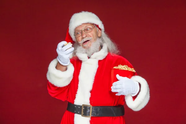 Christmas. Smiling, kind Santa Claus in white gloves with his mouth open holds a red bucket with popcorn with one hand, takes some popcorn and prepares to eat it with the second hand. The concept of — Stock Photo, Image