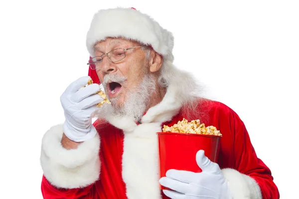 Natale. Sorridente, gentile Babbo Natale in guanti bianchi con la bocca aperta tiene un secchio rosso con popcorn con una mano, prende un po 'di popcorn e si prepara a mangiarlo con la seconda mano. Il concetto di — Foto Stock
