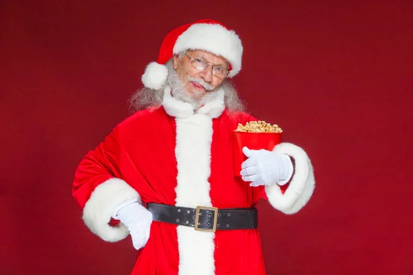 Natale. Sorridente, gentile Babbo Natale in guanti bianchi tiene un secchio rosso con popcorn con una mano, e mette l'altra mano sulla cintura. Il concetto di visitare il cinema, guardare un film con — Foto Stock