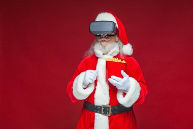 Noel. Noel Baba kırmızı bir kova patlamış mısır tutarak siyah sanal gerçeklik bardaklarda. Sanal gerçeklik bir film izlerken. Kırmızı zemin üzerine izole.