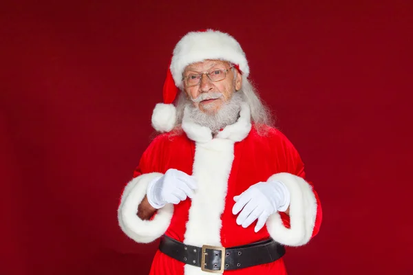 Christmas. Serious Santa Claus in white gloves adjusts her glasses and stares into the camera. Isolated on red background. — Stock Photo, Image