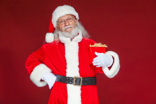 Christmas. Smiling, kind Santa Claus in white gloves holds a red bucket with popcorn with one hand, and puts the other hand on his belt. The concept of visiting the cinema, watching a movie with — Stock Photo, Image