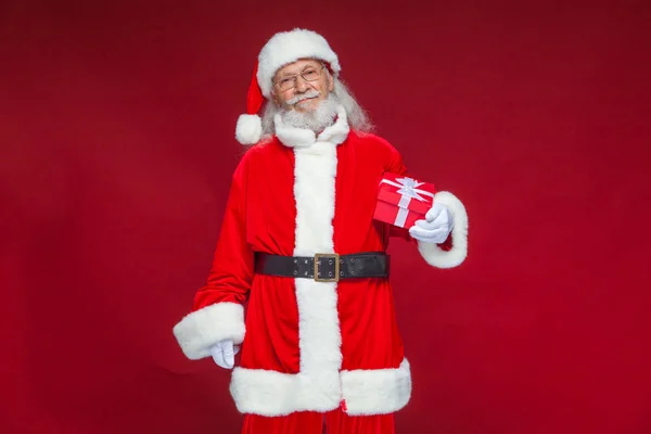 Weihnachten. Lächelnder Weihnachtsmann in weißen Handschuhen hält eine rote Geschenkschachtel mit Schleife in der Hand. zeigt auf das Geschenk. isoliert auf rotem Hintergrund. — Stockfoto