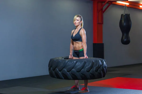 Apto atleta feminino exercitar-se com um pneu enorme, girando e transportar no ginásio. Crossfit mulher exercitando com pneu grande — Fotografia de Stock