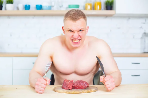 Hombre musculoso con un torso desnudo en la cocina con un trozo de carne de res, el concepto de una dieta saludable. Estilo de vida atlético. Degeneración agresiva depredadora de la cara . — Foto de Stock