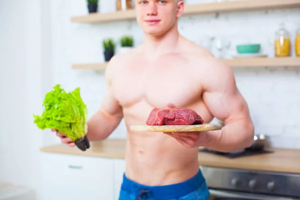 Homem musculoso com um tronco nu na cozinha com uma salada e um pedaço de carne bovina, o conceito de uma dieta saudável. Modo de vida atlético . — Fotografia de Stock