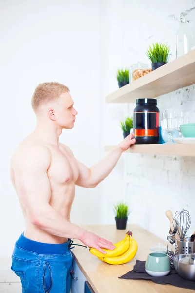 Hombre musculoso con un torso desnudo en la cocina con una proteína nutricional deportiva y plátanos, el concepto de una dieta saludable. Estilo de vida atlético . — Foto de Stock