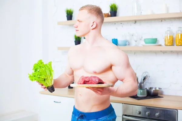 Homem musculoso com um tronco nu na cozinha com uma salada e um pedaço de carne bovina, o conceito de uma dieta saudável. Modo de vida atlético . — Fotografia de Stock