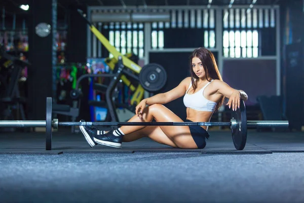 让长腿的女人坐在杠铃附近健身房附近的地板上 — 图库照片