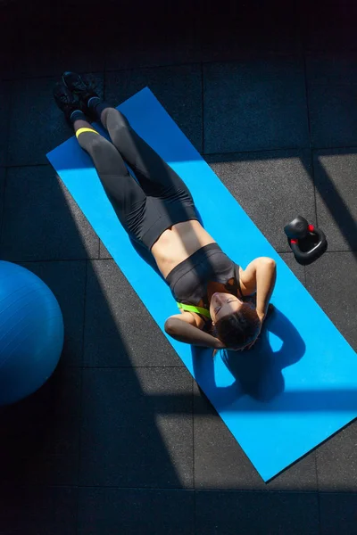 Ajuste a jovem mulher em roupas esportivas deitada no tapete de exercícios perto de fitball e peso fazendo exercícios estomacais. Visão geral do exercício feminino no ginásio — Fotografia de Stock