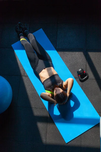 Ajuste a jovem mulher em roupas esportivas deitada no tapete de exercícios perto de fitball e peso fazendo exercícios estomacais. Visão geral do exercício feminino no ginásio — Fotografia de Stock