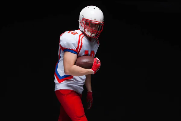 American football player wearing helmet posing with ball on black background