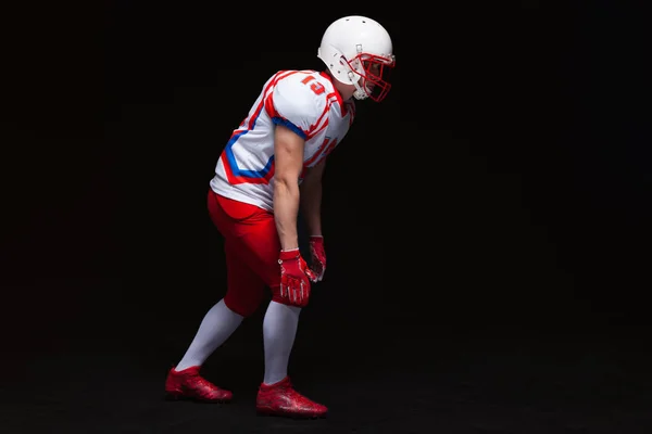 Vista lateral del jugador de fútbol americano que usa casco tomando posición mientras juega contra fondo negro — Foto de Stock