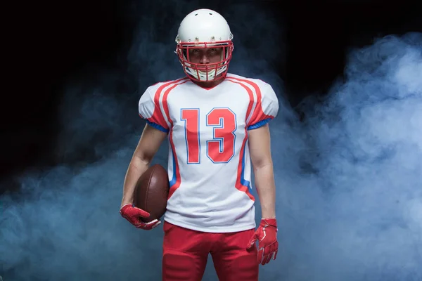 Retrato de vista frontal del jugador de fútbol americano que usa casco con pelota contra humo blanco — Foto de Stock