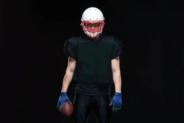 American football player in a black uniform, wearing helmet and holding ball against black background — Stock Photo, Image