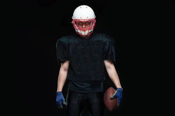 Jugador de fútbol americano con uniforme negro, llevando casco y sosteniendo la pelota sobre fondo negro — Foto de Stock