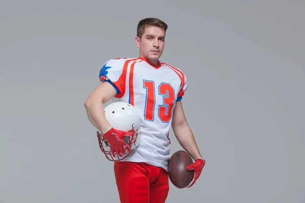 Jugador de fútbol americano sosteniendo pelota de rugby y casco contra fondo gris vista frontal — Foto de Stock