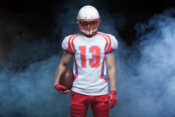 Vorderseite Porträt eines amerikanischen Fußballspielers mit Helm und Ball gegen weißen Rauch — Stockfoto