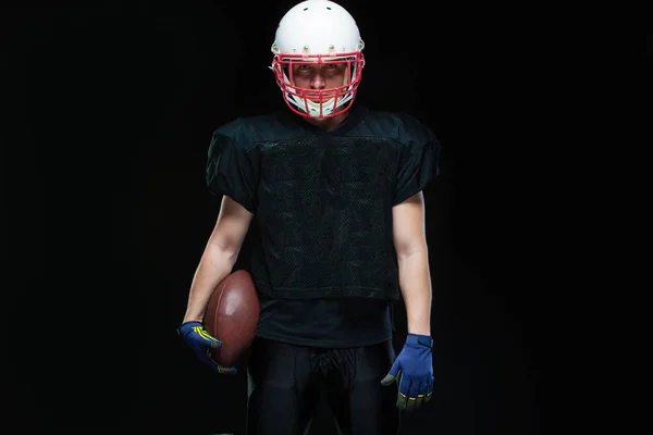 Jugador de fútbol americano con uniforme negro, llevando casco y sosteniendo la pelota sobre fondo negro — Foto de Stock