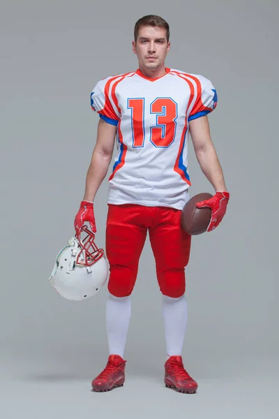 American football player holding rugby ball and helmet against grey background full length Royalty Free Stock Images