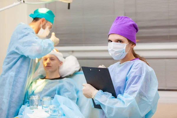 Όμορφο πορτρέτο ενός γιατρού με έγγραφα στο φόντο του λειτουργικού δωματίου. Θεραπεία της φαλάκρα. Μεταμόσχευση μαλλιών. Χειρούργοι στο χειρουργείο διεξάγουν εγχείρηση μεταμόσχευσης μαλλιών. Χειρουργική — Φωτογραφία Αρχείου