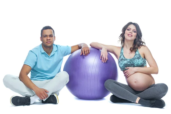 Mladý pár a jejich žena v posledních měsících těhotenství sedí na sportovní rohoži nedaleko míče. Černoch a bílá žena. Koncept šťastného páru různých ras připravujících společně — Stock fotografie