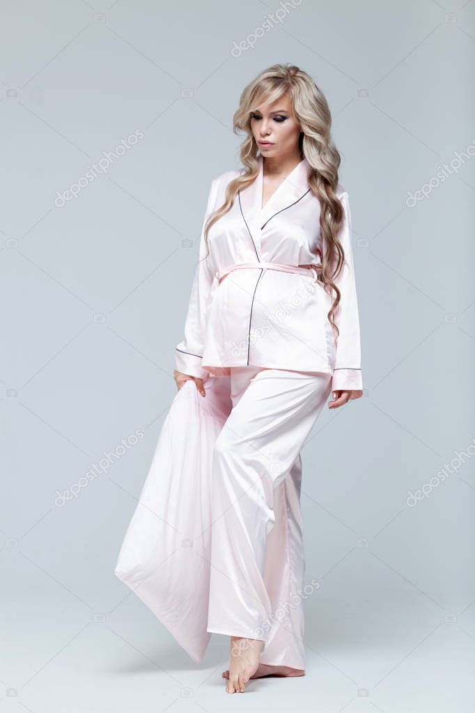 Young beautiful pregnant woman in pink pajamas with pink pillow. The concept of stylish pregnancy and beautiful sleep. On a gray uniform background. Copy paste. Full length.