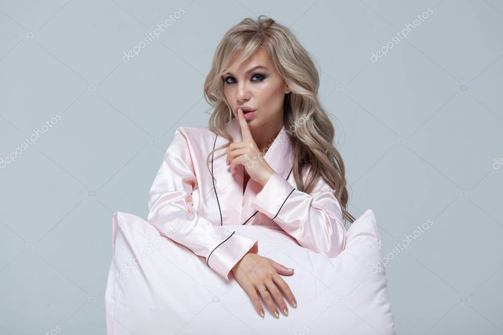 Young beautiful pregnant woman in pink pajamas with pink pillow. Finger to lips, silence. The concept of stylish pregnancy and beautiful sleep. On a gray uniform background. Copy paste.