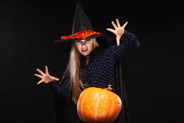 Halloween čarodějka s dýní na černém pozadí vrhá zaklínadlo do rukou. Krásná mladá překvapená žena v čarodějové čepici a kostýmech držící dýni. Široký Halloween umělecký design. Kopírovat-vložit — Stock fotografie