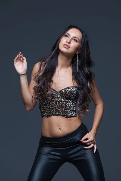 Bella, sexy e sensuale ragazza modello bruna con trucco luminoso in un top di stampa leopardo alla moda e pantaloni di pelle nera seducente guardando la fotocamera e posa su sfondo scuro Foto Stock
