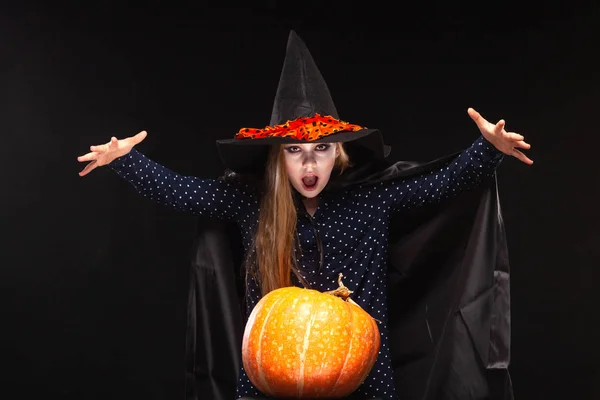 Halloween čarodějka s dýní na černém pozadí vrhá zaklínadlo do rukou. Krásná mladá překvapená žena v čarodějové čepici a kostýmech držící dýni. Široký Halloween umělecký design. Kopírovat-vložit — Stock fotografie
