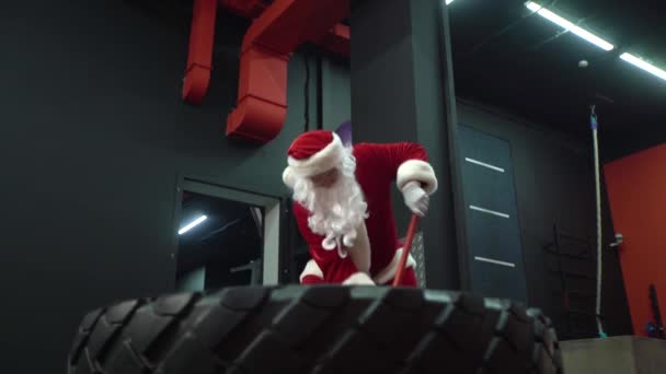 Santa Claus entrenando en el gimnasio el día de Navidad. Santa Claus golpeando neumático de la rueda con el martillo . — Vídeos de Stock