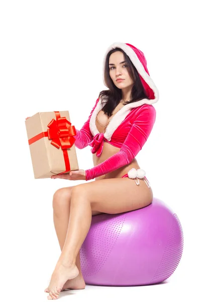 Navidad. Chica deportiva fitness en un traje rojo sexy de Santa Claus sosteniendo una caja de regalo atada con una cinta roja con un lazo y sentado en una pelota de fitness. Aislado sobre fondo blanco . — Foto de Stock